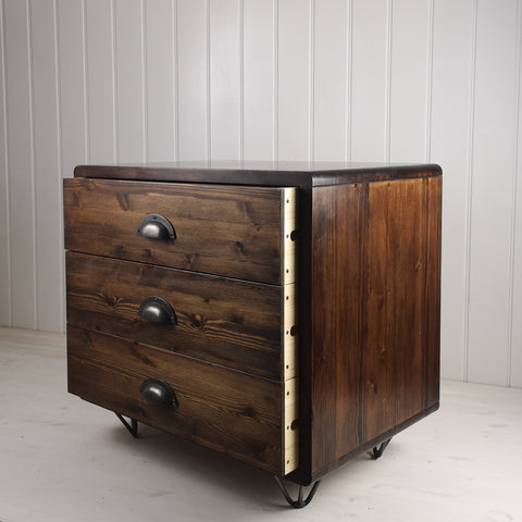 Office furniture, bedside table or side table with three deep drawers. Stained in a dark wood finish with contrasting light wood inside the drawers. Cast iron cup handles and standing on retro hairpin feet.